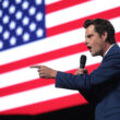 Matt Gaetz speaking into a microphone in front of a video screen showing an American flag.