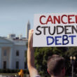 White young man with his back to the viewer is holding a hand made sign that reads, "Cancel student debt."