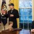 Elon Musk, in a long black coat and black MAGA hat, takes questions from reporters as Donald Trump looks on from the Resolute desk in the Oval Office.