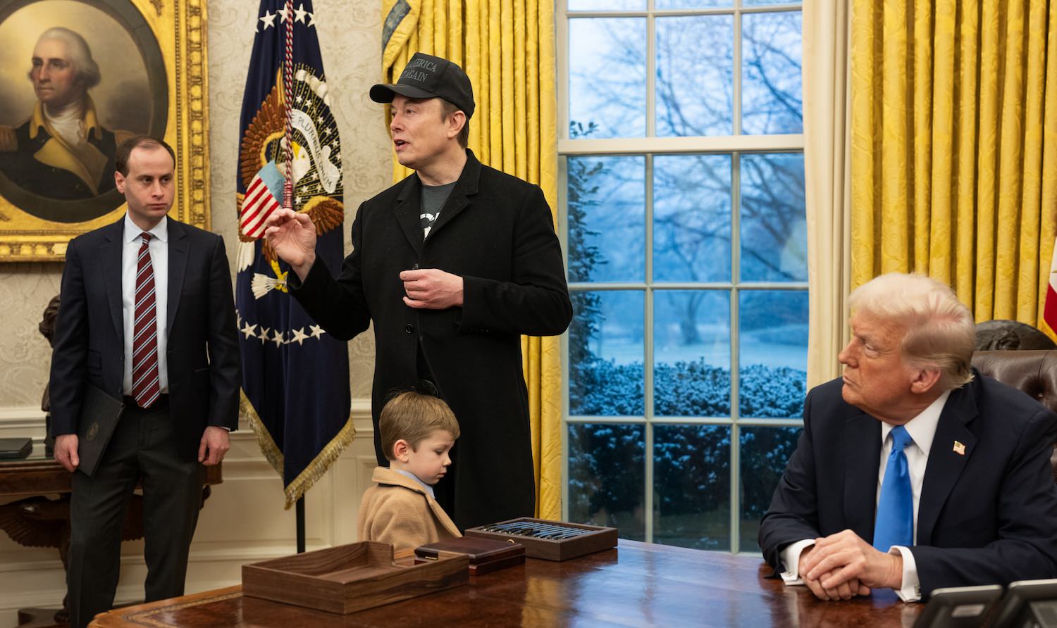 Elon Musk, in a long black coat and black MAGA hat, takes questions from reporters as Donald Trump looks on from the Resolute desk in the Oval Office.