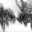 Beach activity at Da Nang, Vietnam during landing of United States Marines of the 9th Marine Expeditionary Brigade in March of 1965. A group of U.S. soldiers are marching towards the camera in a grainy black and white photo.
