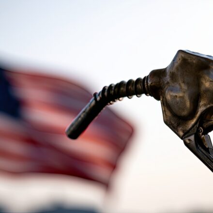 A close-up of a gasoline pump nozzle in focus, with a blurred American flag in the background, representing fuel costs and the economy.