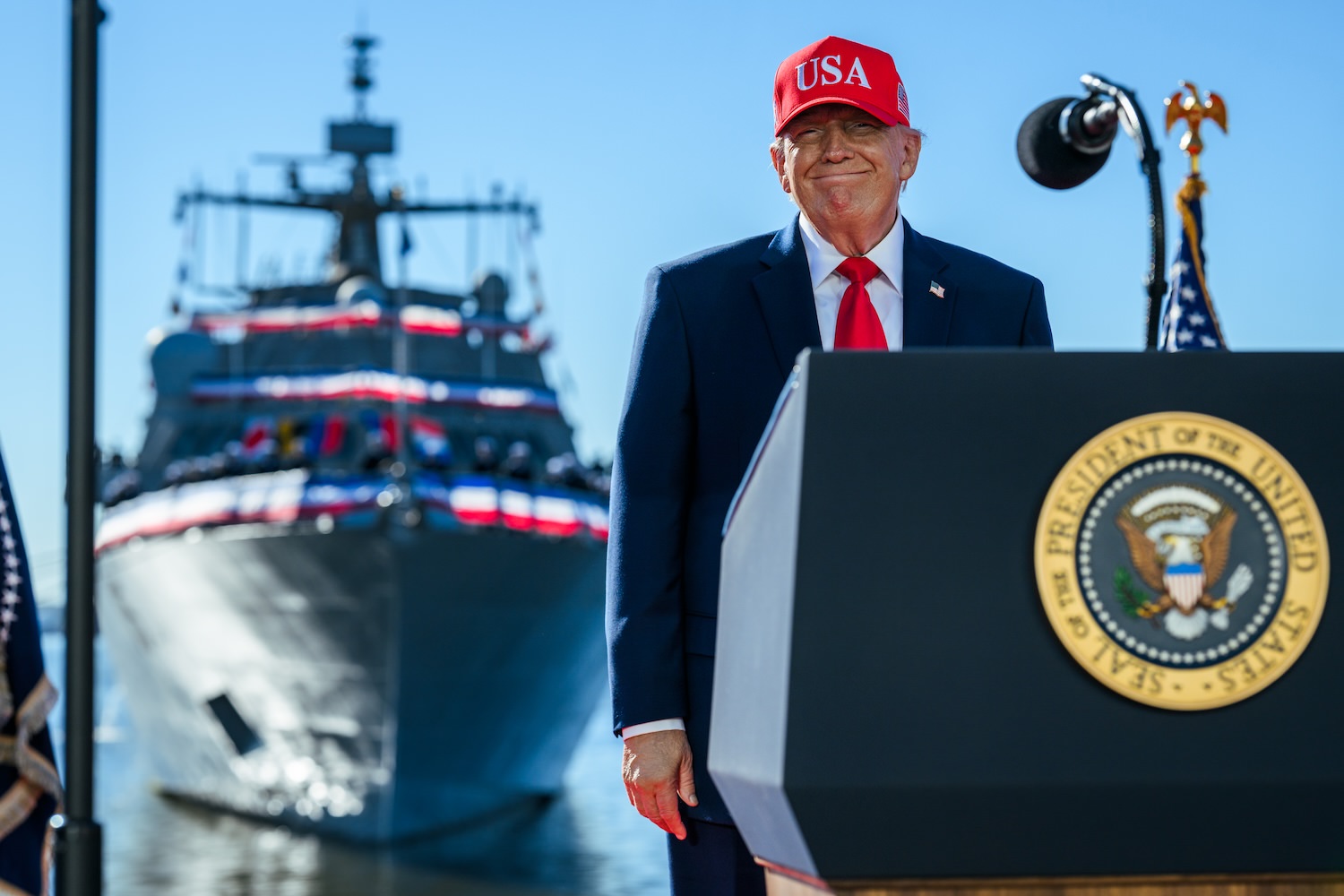President Donald J. Trump standing and smiling with a red USA baseball cap on behind the Presidential podium. At Sparrows Point, Baltimore, Maryland, Tuesday, October 6, 2026. (Official White House Photo by Daniel Torok)
